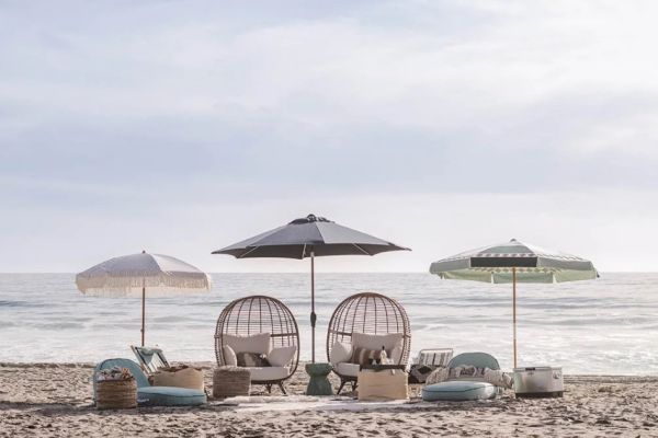 Beach setup with umbrellas, egg chairs, cushions, and decor facing the ocean under a cloudy sky.