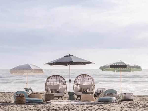 Beach setup with umbrellas, egg chairs, cushions, and decor facing the ocean under a cloudy sky.