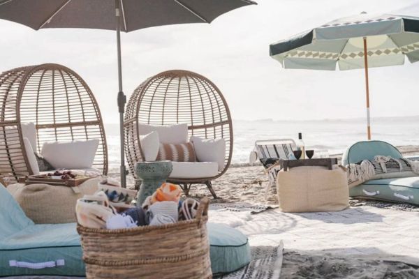 The image shows a beach setup with wicker chairs, umbrellas, and cushions on sand, creating a cozy and relaxed atmosphere.