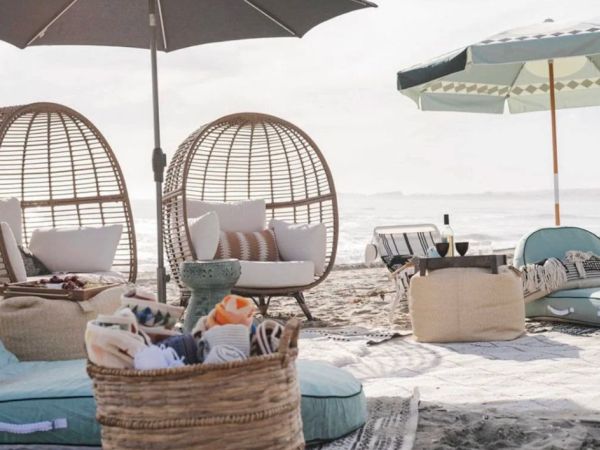 The image shows a beach setup with wicker chairs, umbrellas, and cushions on sand, creating a cozy and relaxed atmosphere.