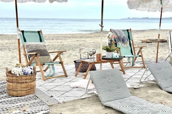Beach scene with loungers, umbrellas, and a picnic setup including blankets, pillows, and drinks near the calm sea.