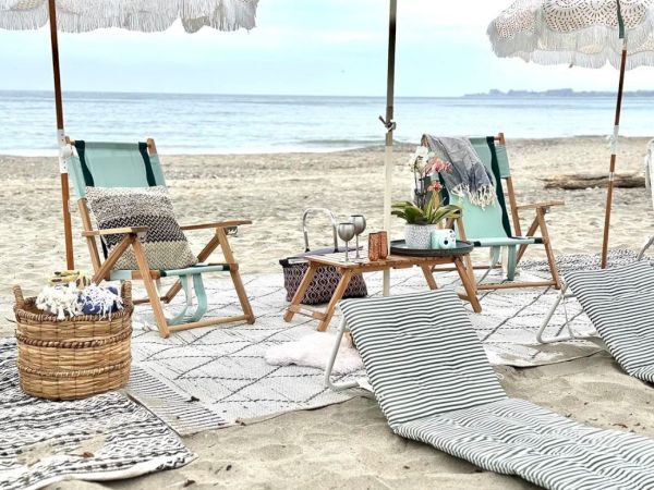 Beach scene with loungers, umbrellas, and a picnic setup including blankets, pillows, and drinks near the calm sea.