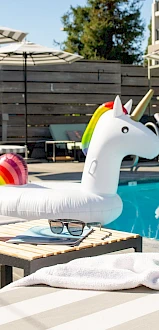 A poolside scene with a unicorn float, beach ball, and a cocktail. Lounge chairs and umbrellas in the background complete the sunny setting.