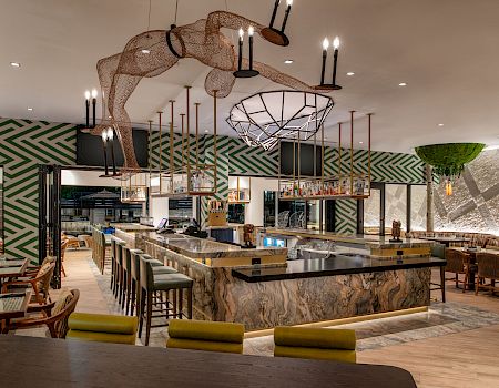 A stylish restaurant interior with a long central bar, geometric green walls, modern lighting, and various seating areas around.