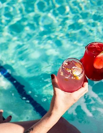 Two people toast cocktails by a pool, sunlit blue water, one with a black straw, green bikini visible, tattoos on arm, cheerful vibe.