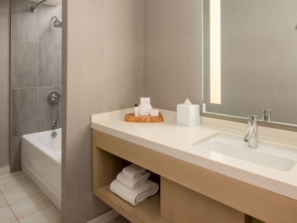 A modern bathroom with a vanity sink, toiletries, bright mirror light, tiled shower, and a toilet in the corner.
