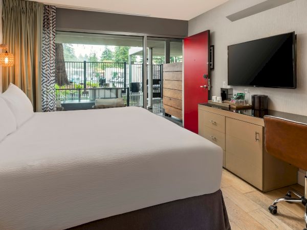 A hotel suite shows a large bed, curtains, a balcony view, and a TV on a cabinet near a desk and a red door.