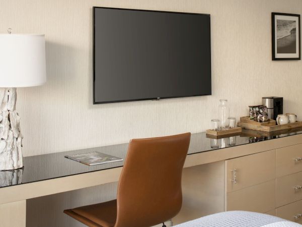 Modern hotel room with a wall-mounted TV, desk lamp, framed photos, glass jars and a dresser beside a chair.