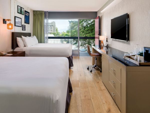 A modern hotel room with twin beds, a desk, wall art, pendant lights, and a large window showing greenery outside.