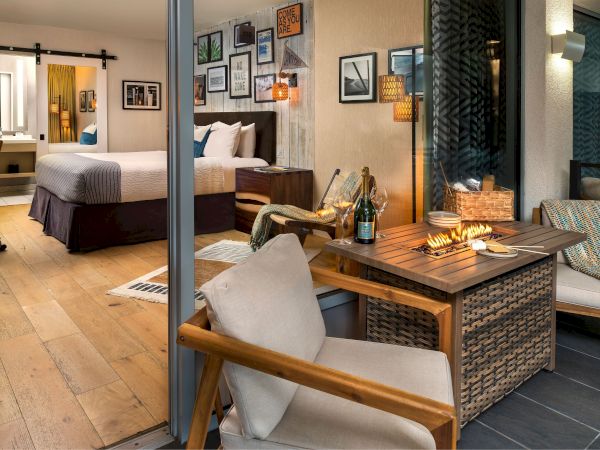 A cozy suite patio with a lit grill, wooden table set for drinks, wicker chairs, and a glimpse of a bed inside.