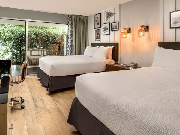 A modern hotel room with two beds, warm wall lamps, framed art, and a balcony door opening to greenery.