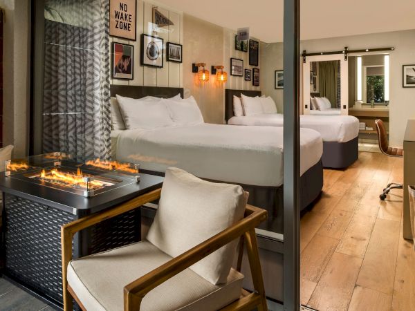 A stylish hotel suite with a flame firepit coffee table, twin beds, greenery-dark curtains, and framed art, plus a &ldquo;NO WAKE ZONE&rdquo; sign.