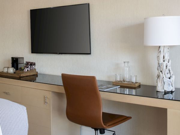 A hotel-style room showing a desk with glassware, a lit lamp, and a wall-mounted flat-screen TV.