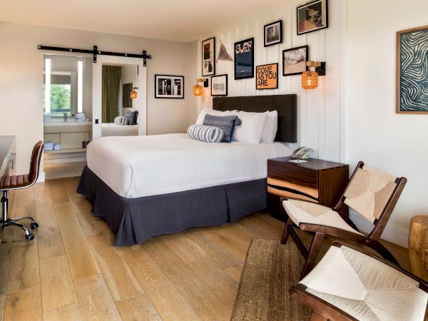 A modern hotel bedroom with a large bed, pillows, framed wall art, a desk and chair, and warm pendant lights.