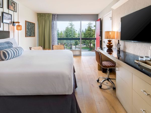 A modern hotel room with a king bed, patterned curtains, and a desk area with TV and window views of trees.