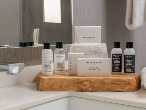 A bathroom sink with a wooden tray holding Acca Kappa bottles, soap, and rolled towels near a shower curtain.