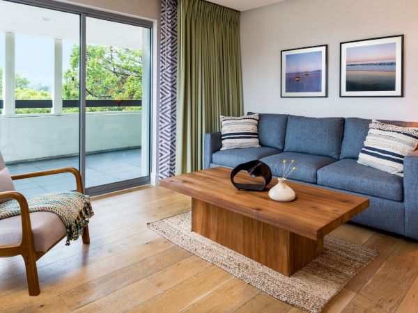 A cozy living room with blue sofa, wooden coffee table, accent chairs, woven lamps, wall art, and large balcony doors.