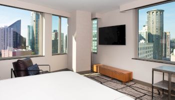 This image shows a modern hotel room with large windows offering a city view, a wall-mounted TV, a bed, an armchair, and a small table.