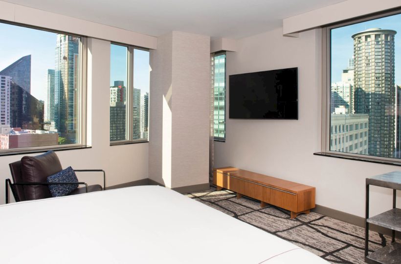 This image shows a modern hotel room with large windows offering a city view, a wall-mounted TV, a bed, an armchair, and a small table.