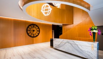 A modern lobby with a reception desk, a large wall clock, spiral wooden design, white marble floors, and decorative lighting fixtures on the ceiling.