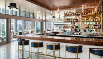 This image shows a modern bar with a wooden countertop, five blue bar stools, and shelves stocked with various bottles behind the counter.