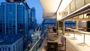 The image shows a modern balcony view at dusk, overlooking a cityscape with tall buildings and a street below, featuring seating and tables.