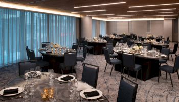 The image shows a banquet hall with round tables set for an event, complete with elegant tableware, black napkins, and flower centerpieces.