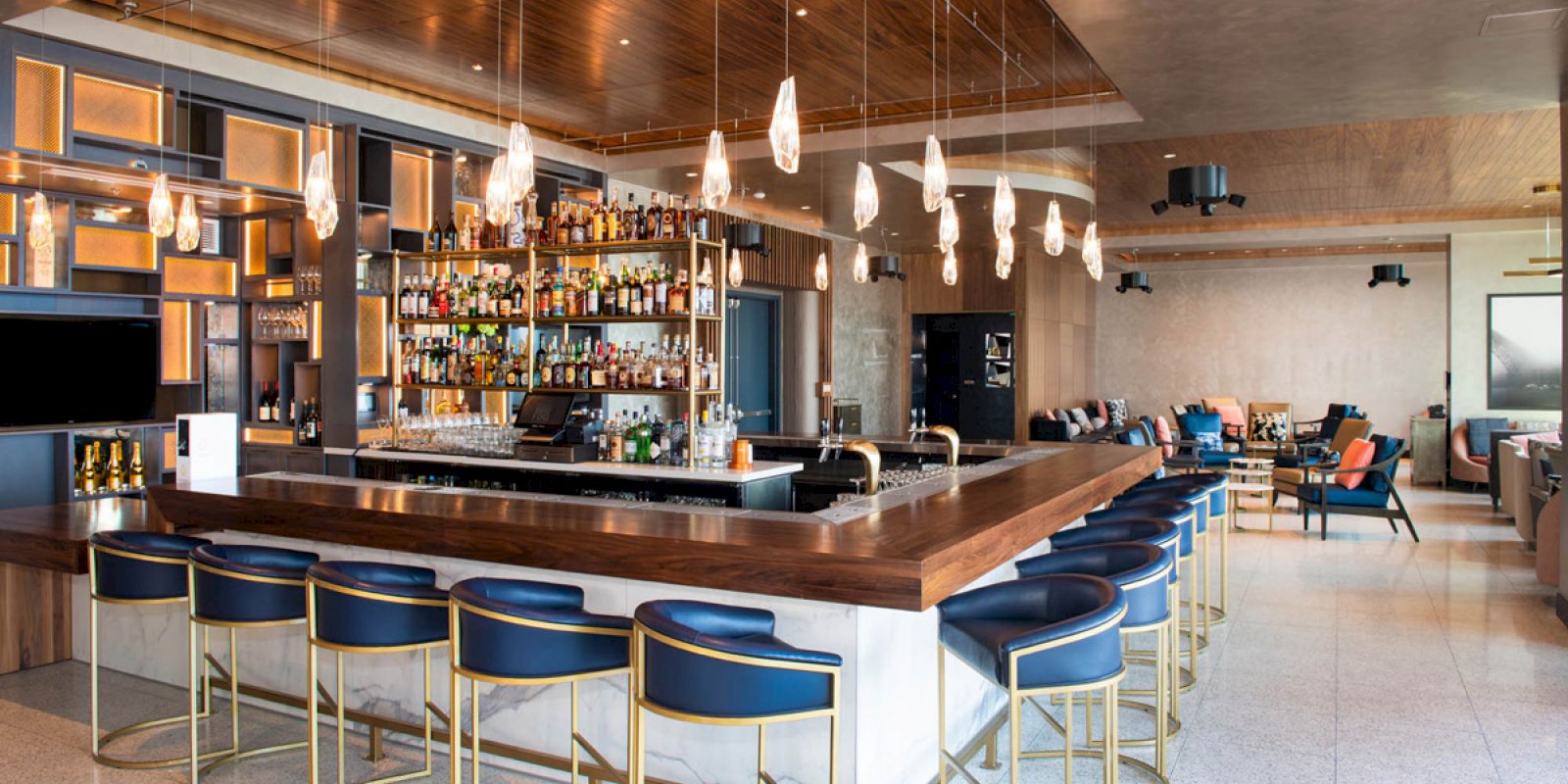 A modern bar with blue stools, a liquor display, pendant lights, and a lounge area with cozy seating arrangements in a stylish interior design.