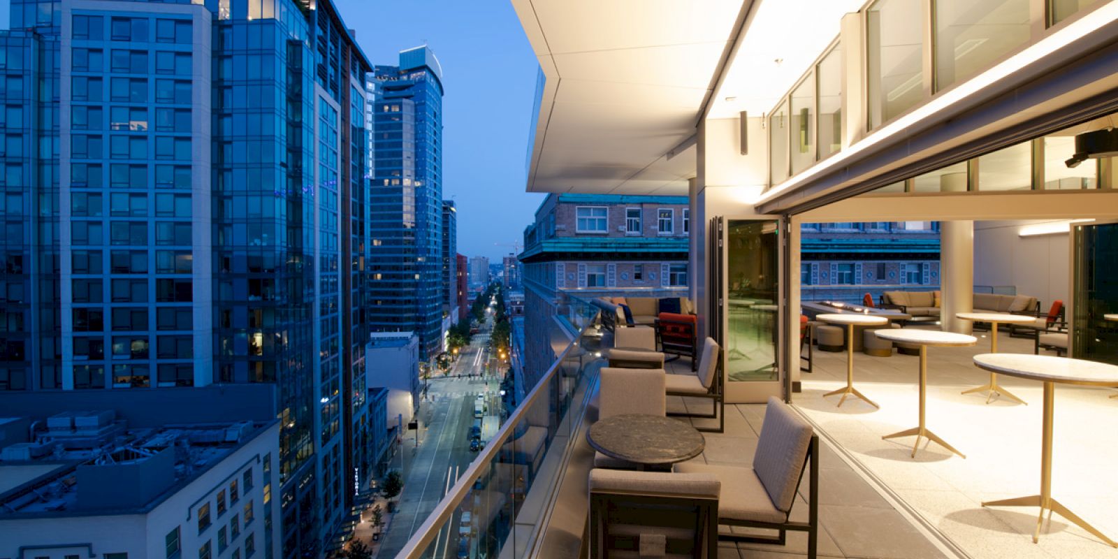 A modern city balcony view featuring outdoor seating with tables and chairs, overlooking lit-up tall buildings and a street below in the evening.