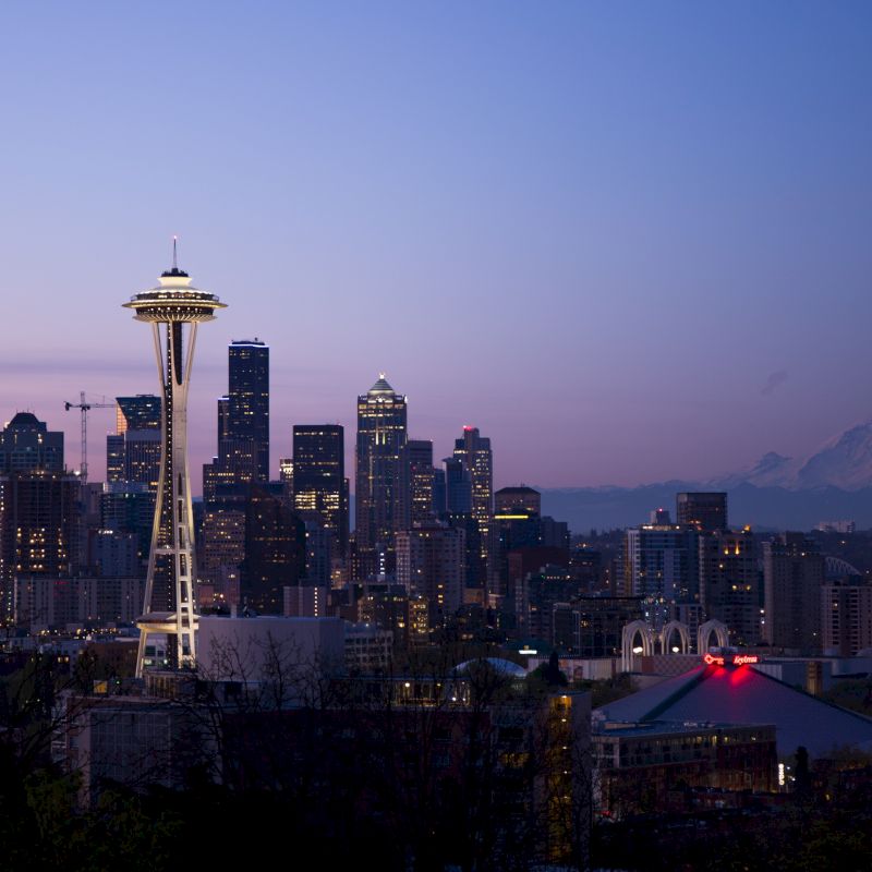 A nighttime skyline photo of Seattle, featuring the Space Needle and Mount Rainier in the background, with city lights illuminating the scene.