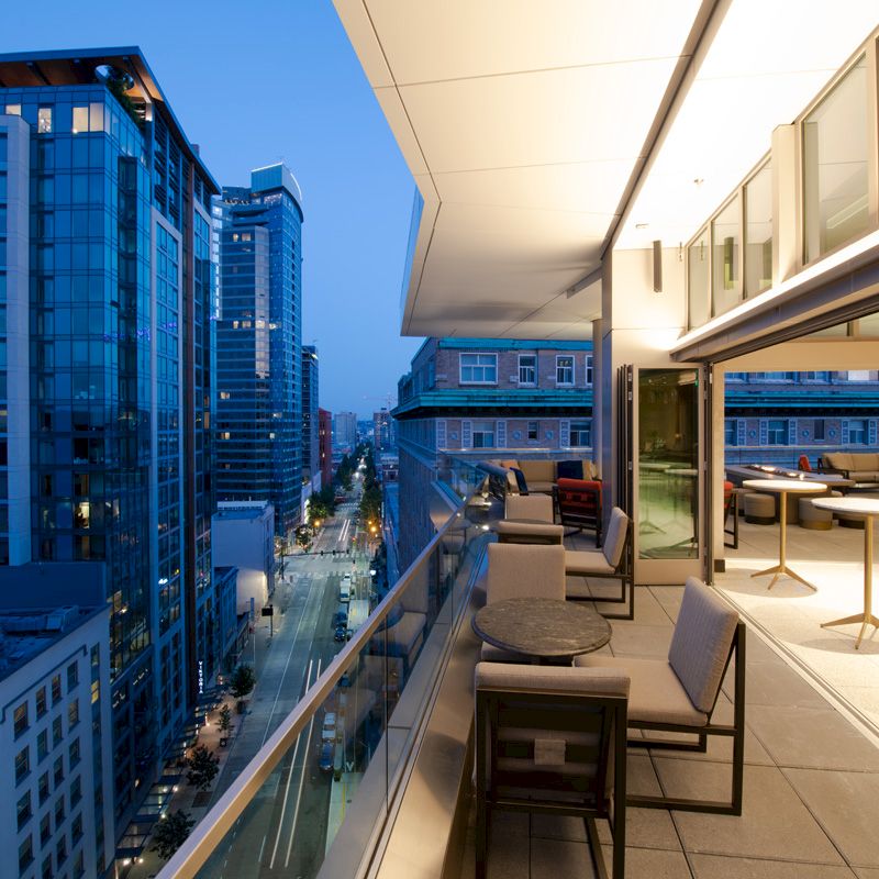 The image shows a modern cityscape at dusk, with tall buildings lining a street, and a rooftop terrace with outdoor seating and tables.