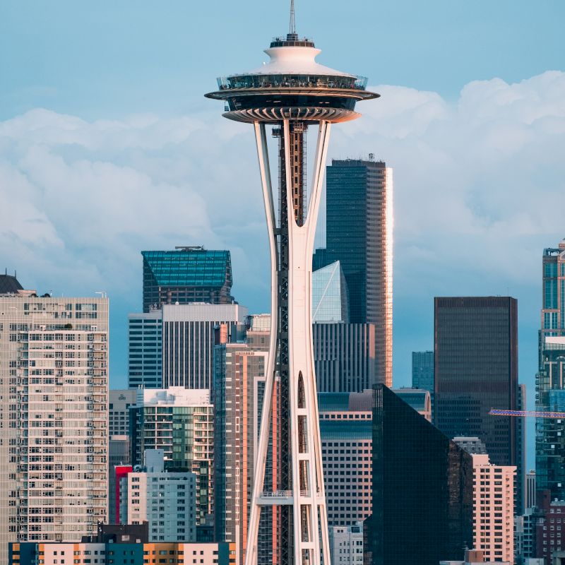 The image shows the Space Needle, an iconic tower set against a background of modern skyscrapers and buildings, with a cloudy sky in Seattle.