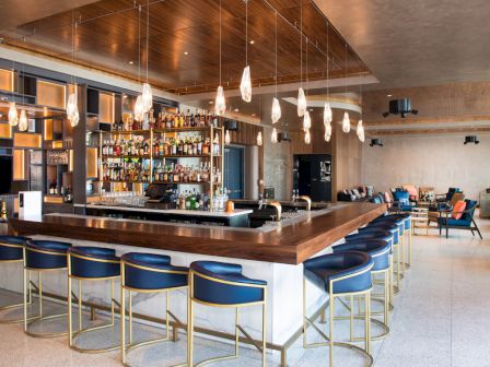 The image shows a modern bar with blue stools, stylish lighting, and a well-stocked bar in the center, with seating areas in the background.