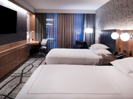 This image shows a modern hotel room with two beds, a TV on the wall, a desk, and a chair by the windows, with clean and stylish decor.