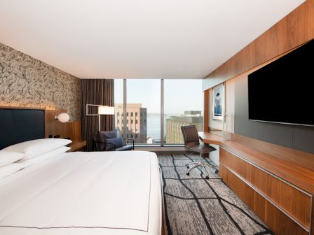 A modern hotel room features a large bed, flat-screen TV, desk, and panoramic windows with a city view.