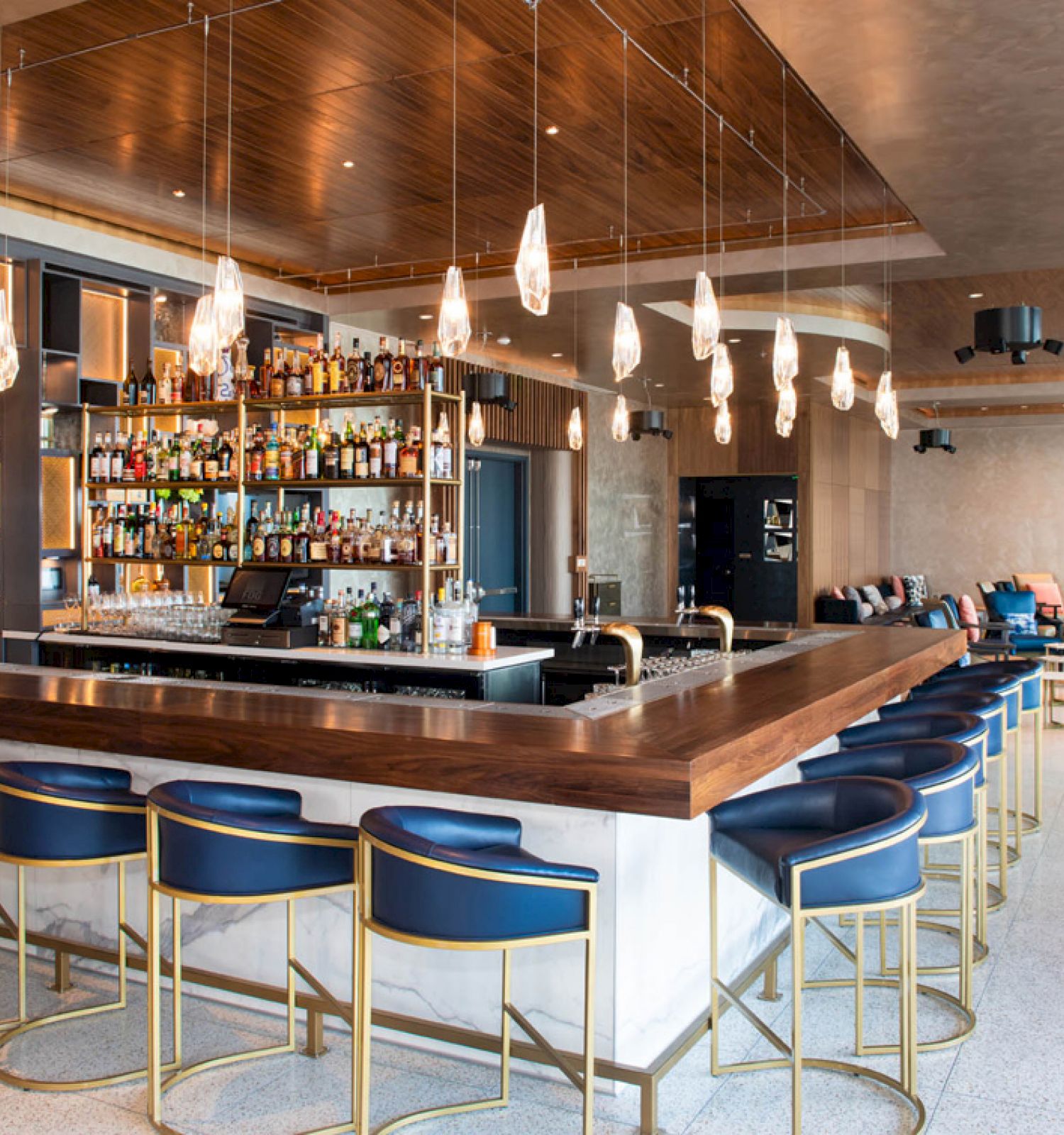 The image shows a modern bar with blue chairs, a central bar counter, shelves of liquor bottles, hanging pendant lights, and a seating area.