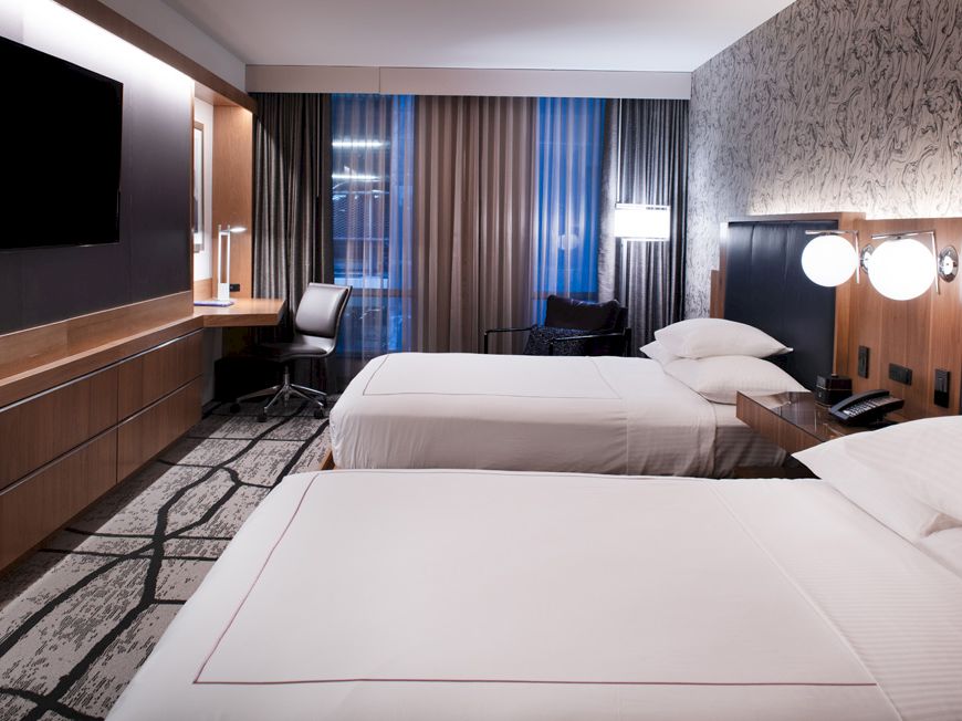 A modern hotel room with two beds, a wall-mounted TV, a desk with a chair, and stylish decor featuring patterned wallpaper.