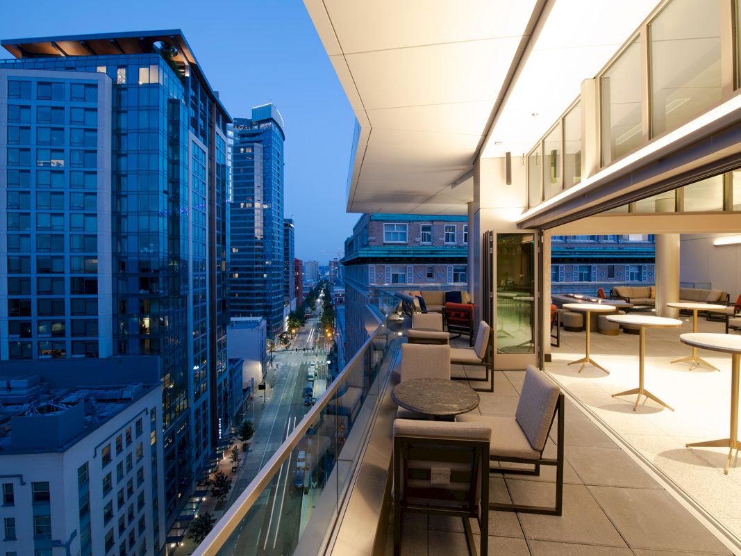 The image shows a modern cityscape during twilight, viewed from a balcony with tables and chairs, overlooking high-rise buildings and a street below.