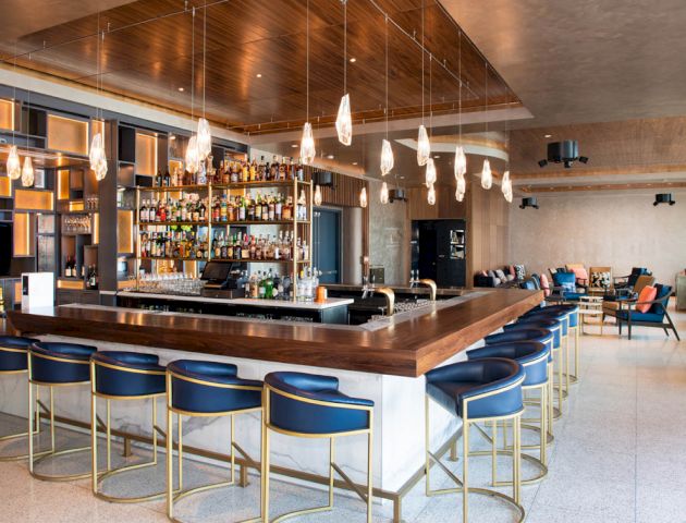 This image shows a modern bar with a central counter, blue bar stools, hanging lights, and a fully stocked bar. Seating areas are visible in the background.