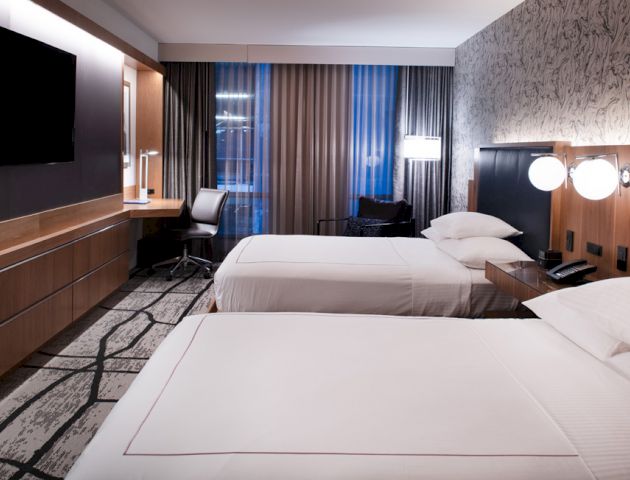 A modern hotel room with two neatly made beds, a wall-mounted TV, a desk with a chair, and night lamps on the headboard, softly lit.