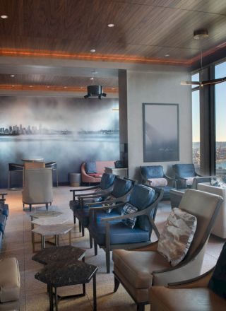 A modern lounge area with comfortable chairs, tables, large windows offering a view of a waterfront cityscape including a Ferris wheel.