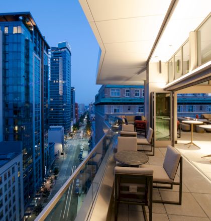 A modern cityscape at dusk featuring high-rise buildings and a well-furnished balcony with tables, chairs, and a view of the street below.