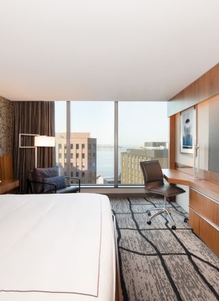 A modern hotel room with a large bed, wall-mounted TV, work desk, and city view through floor-to-ceiling windows.