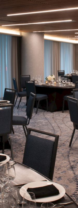 A modern banquet hall is set with round tables, chairs, and elegant place settings under warm lighting. The room has a calm, sophisticated ambiance.