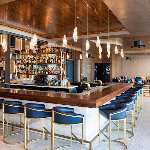 This image shows a modern bar with a wooden countertop, blue stools, and hanging lights. The back has shelves filled with liquor bottles and glasses.