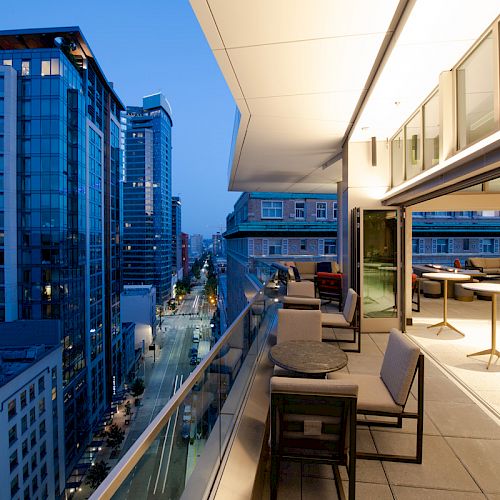 A modern balcony with seating areas overlooks a cityscape with tall buildings at dusk, offering an urban view of the surrounding architecture.