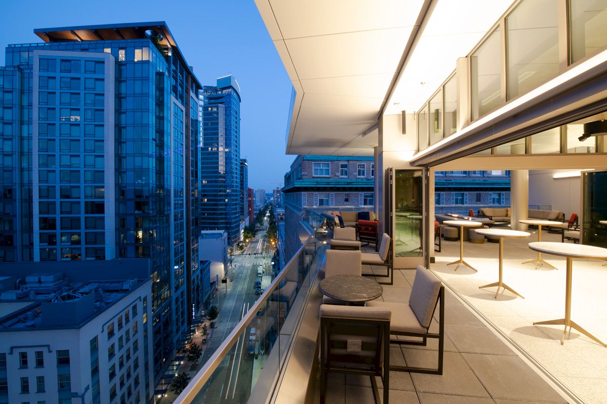 A modern outdoor balcony with seating and tables, overlooking a city street lined with tall buildings in the evening.