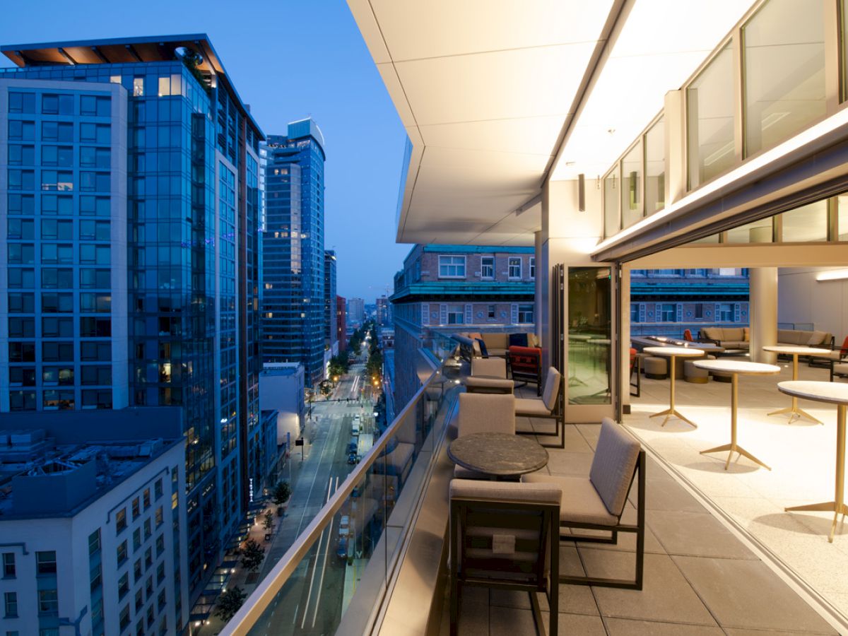 A modern outdoor balcony with seating and tables, overlooking a city street lined with tall buildings in the evening.