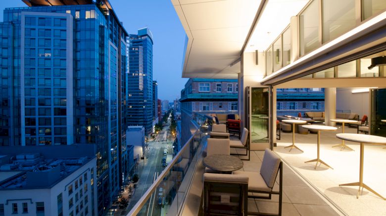 A modern outdoor balcony with seating and tables, overlooking a city street lined with tall buildings in the evening.