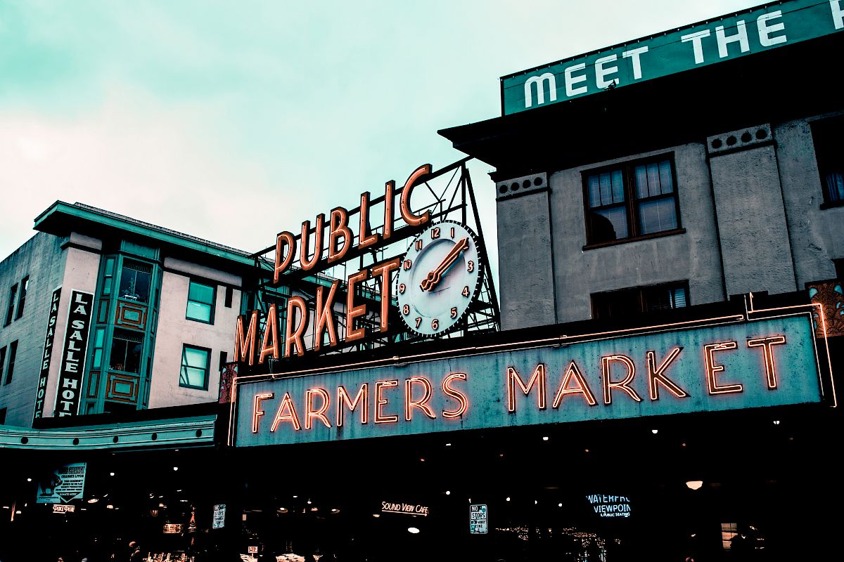 The image shows a neon sign for a "PUBLIC MARKET" and "FARMERS MARKET" with buildings in the background, possibly in a busy urban area.